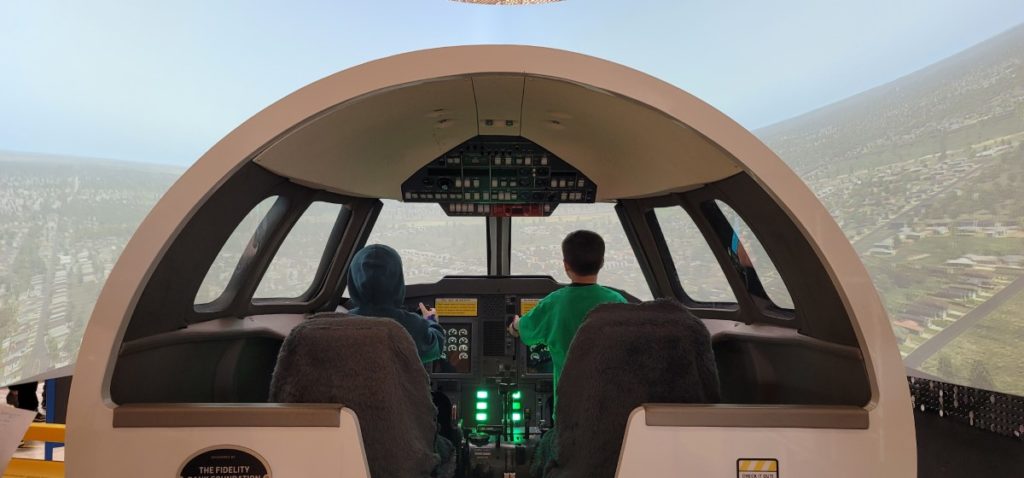 Two kids playing in an airplane cockpit in front of a screen with a picture of the sky.