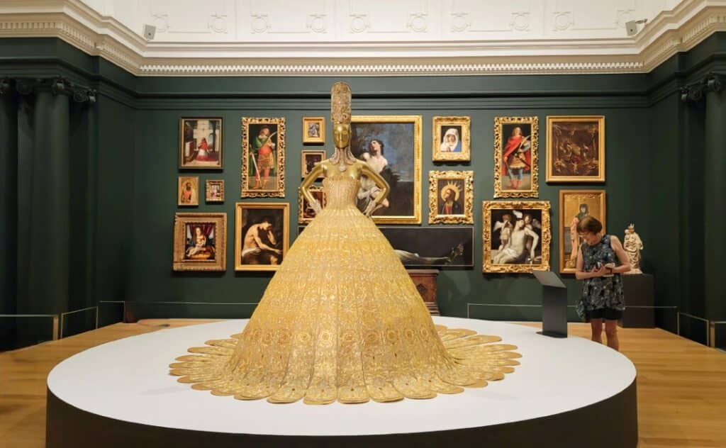 A bright gold dress on a statue with art pieces hanging behind it at the Auckland Gallery Toi o Tamaki.